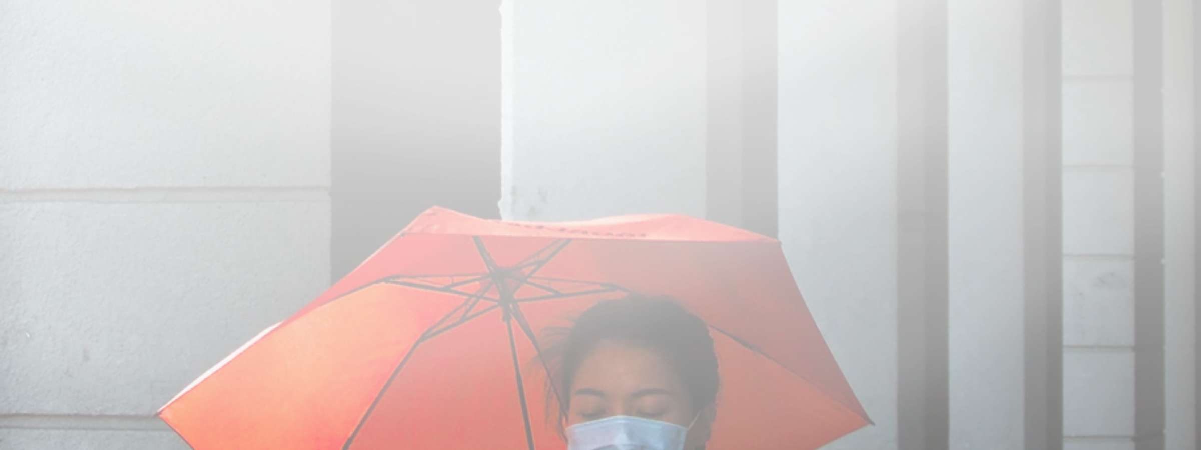 A woman wearing a face mask and carrying a red umbrella walks down a rainy street, while looking at her phone